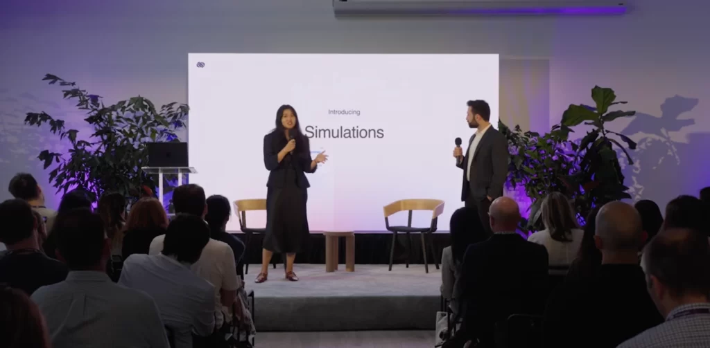 Two presenters speaking in front of a large event screen with seated attendees.