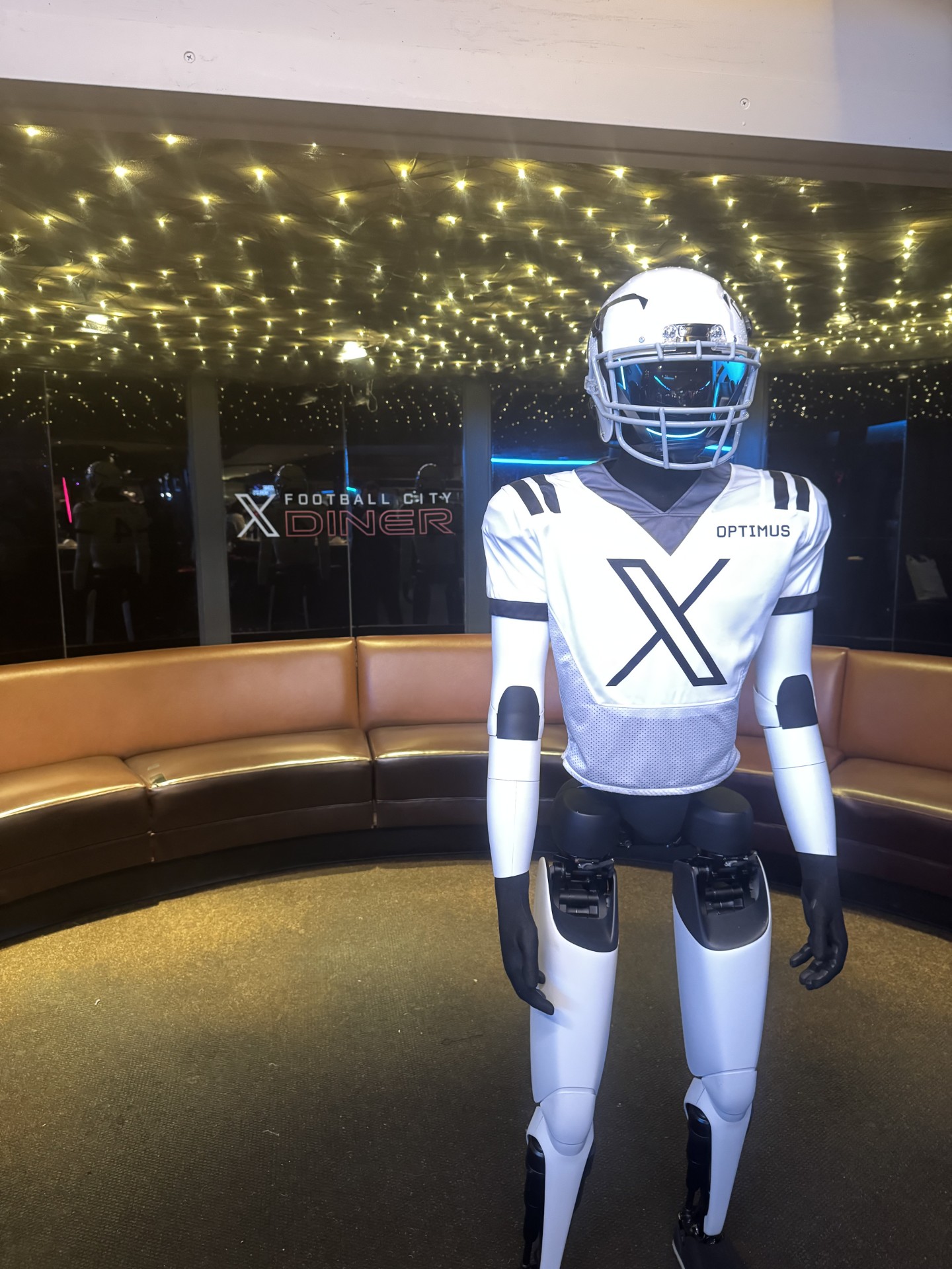 An Optimus robot in an X jersey standing under the twinkly light ceiling at X Football City Diner, with the curved banquette behind