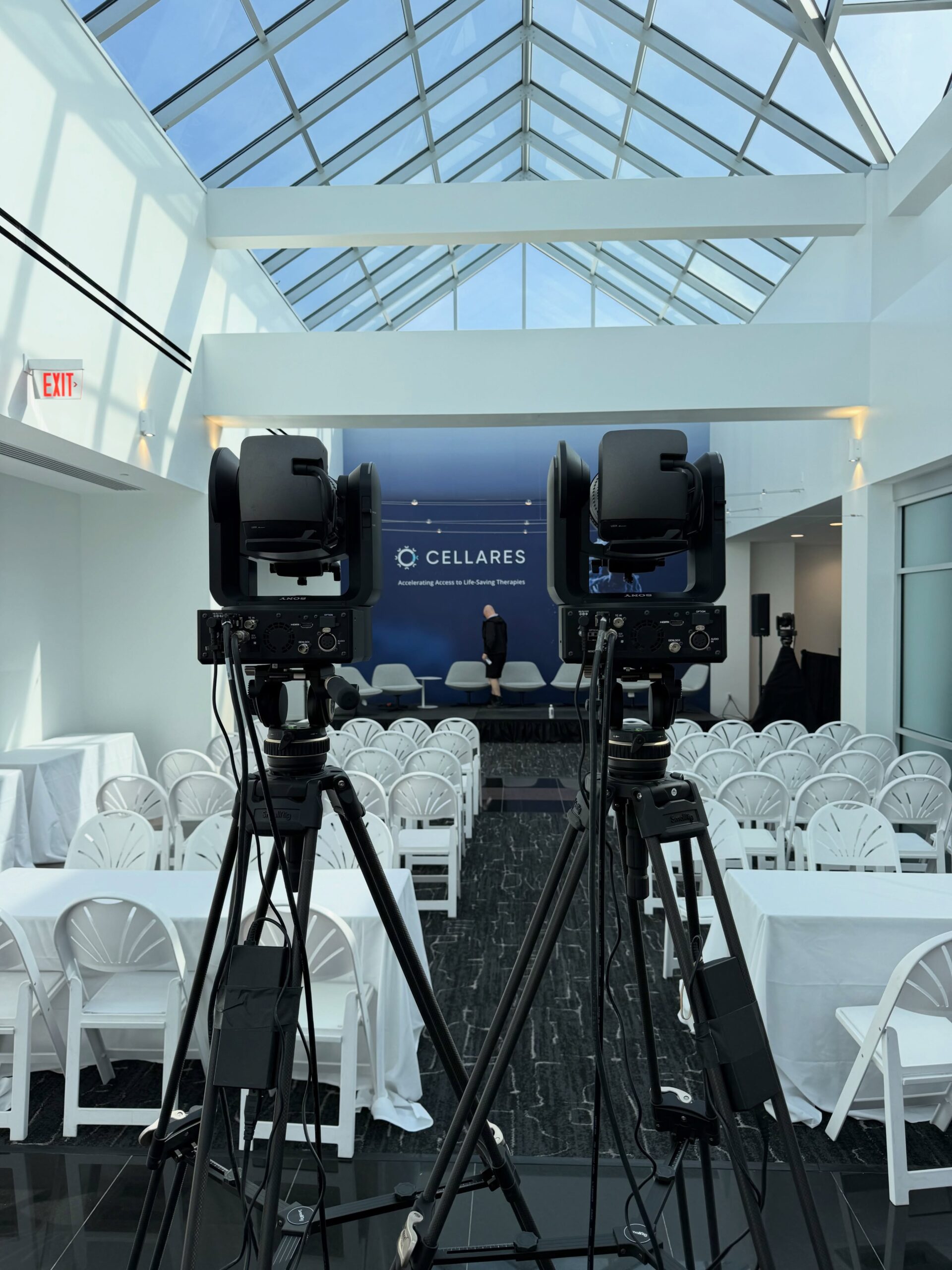 Two broadcast cameras on tripods facing a Cellares-branded panel set under a glass atrium roof