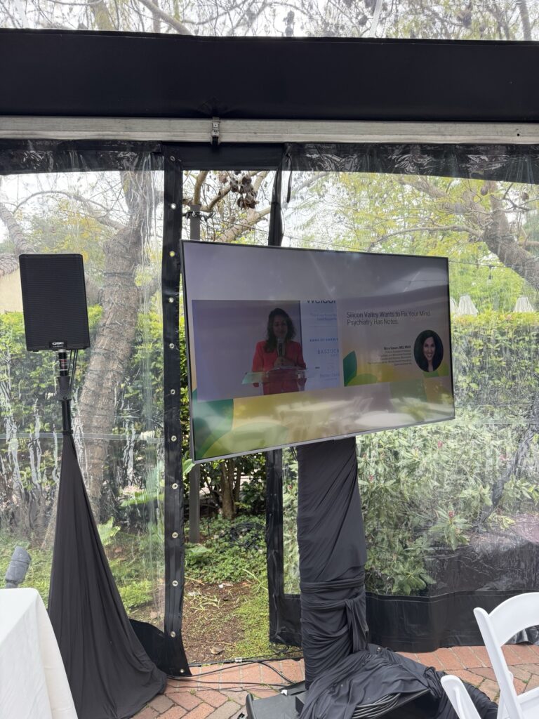 Outside tent at Allied Arts Guild with a 65 inch screen and speaker on stands carrying the live camera feed from the main session