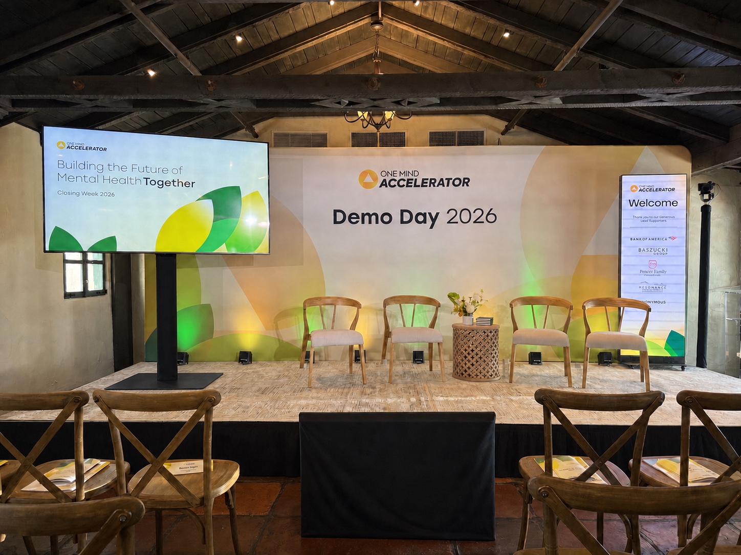 Demo Day 2026 main session stage at Allied Arts Guild, with printed backdrop, side screen on a black floor-base stand, and stage lighting set by Westwave AV