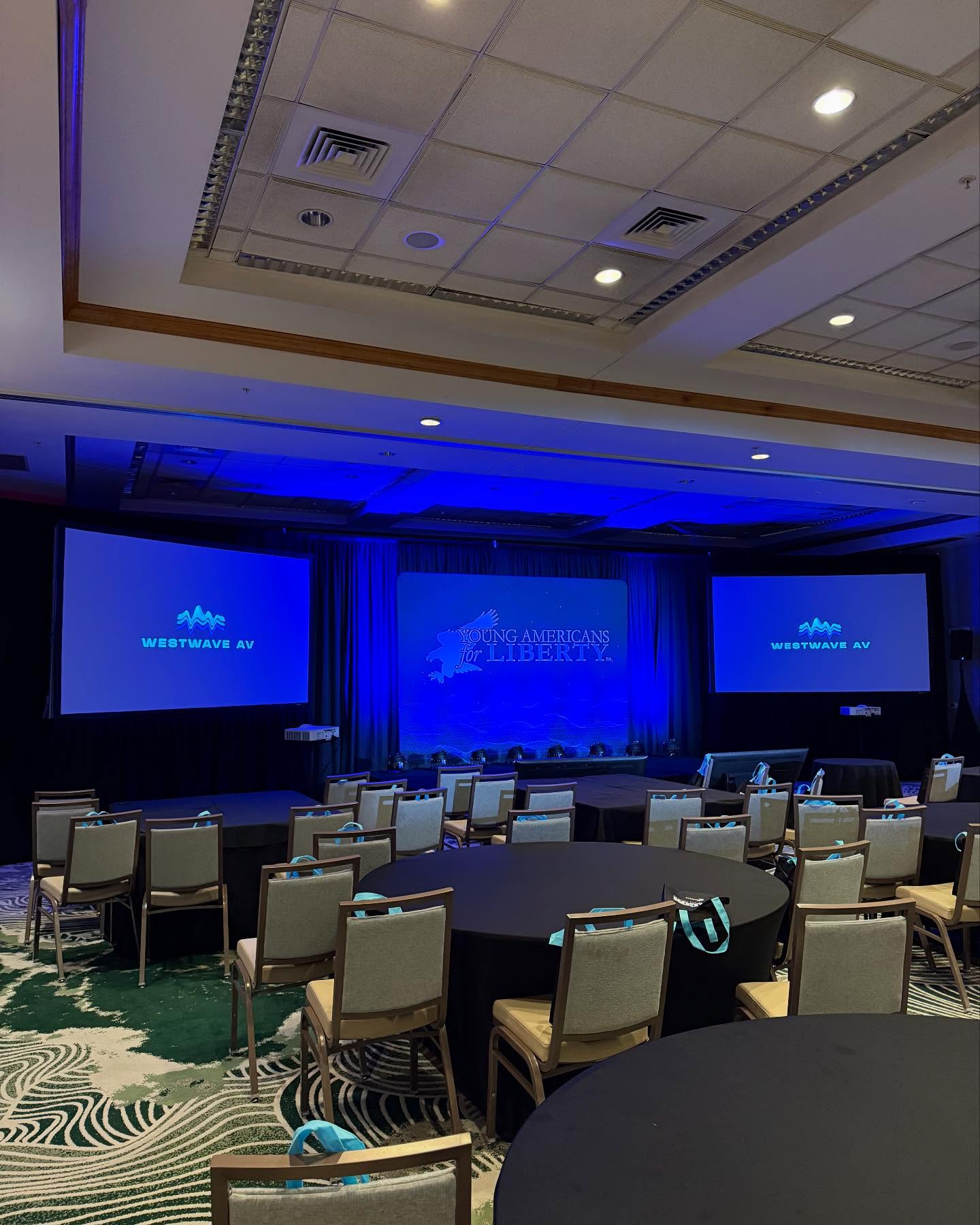 A hotel ballroom in a deep blue wash with three screens carrying a Westwave AV holding slide and round tables set